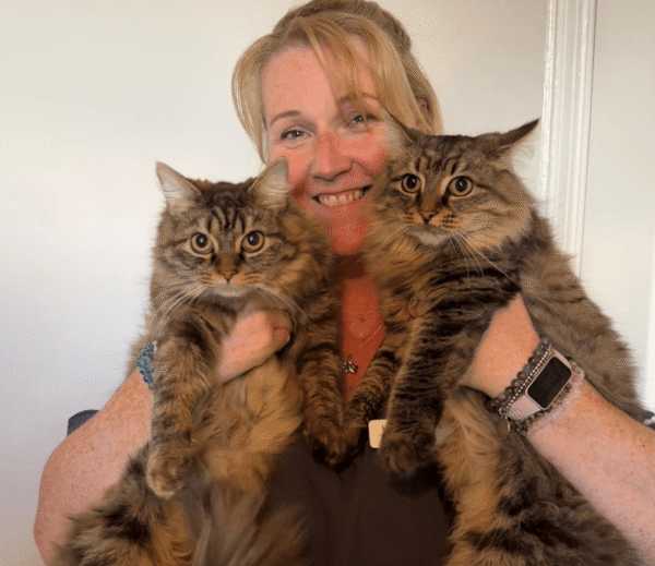 Receptionist Khadine Rich smiling while holding two fluffy tabby cats, both looking directly at the camera indoors against a plain white background.