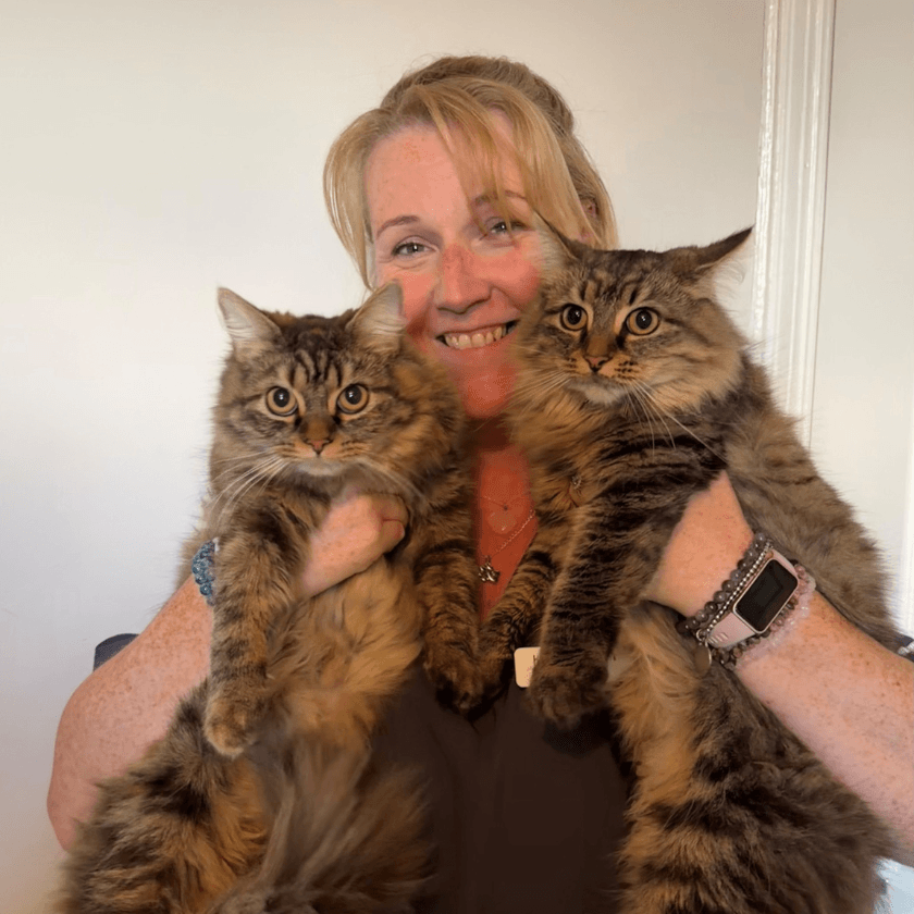 Receptionist Khadine Rich smiling while holding two fluffy tabby cats, both looking directly at the camera indoors against a plain white background.